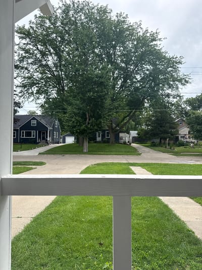 The image captures a view from a white porch railing, looking out onto a suburban street lined with trees and houses. In the foreground, a neatly manicured green lawn is bordered by a concrete walkway leading away from the viewer. Beyond the lawn, a paved driveway cuts through the grass, leading to a dark blue house with white trim and windows. Adjacent to the house is a white garage with a single garage door. Further down the street, other houses and trees are visible under a cloudy sky. The overall impression is of a quiet, residential neighborhood. The location is not specified in the image or the prompt.