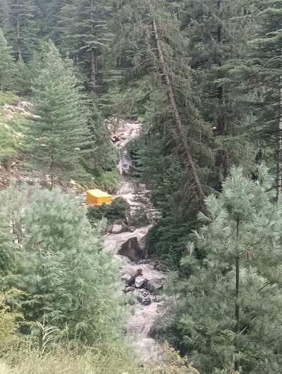 The image shows a dense forest with a stream flowing through it. A small yellow structure, possibly a tent or a shelter, is visible on the bank of the stream. The trees are tall and green, suggesting a natural landscape. The stream appears to be flowing rapidly over rocks, with some white water visible. The overall impression is one of a remote, natural setting within the city of Khakhnal, India, given the context of the provided prompt.