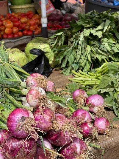 The image captures a close-up view of a produce stall, likely in Mbar, Nigeria, showcasing a vibrant assortment of fresh vegetables. In the foreground, a pile of red onions with their roots still attached are prominently displayed. Behind them, bunches of green onions and leafy greens are arranged, suggesting a bustling market environment. In the background, red tomatoes are visible, along with what appear to be cabbages. The overall impression is one of abundance and freshness, characteristic of a local market. There are no people visible in the frame, but the presence of flies suggests the food is fresh and outdoors. The lighting suggests it is daytime.