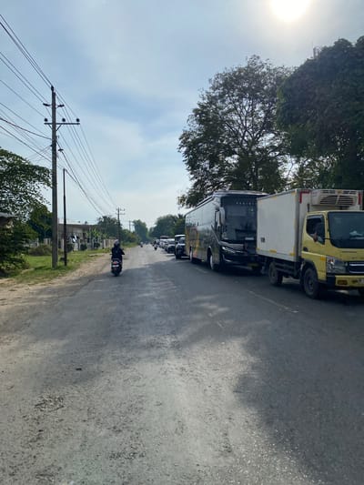 This media file depicts a street scene in Meuse, Indonesia, under a bright, possibly hazy, daytime sky. A line of vehicles, including a yellow refrigerated truck and a large bus, are stopped on an asphalt road. A person on a dark-colored scooter rides past the stationary traffic. Power lines stretch across the frame, supported by wooden poles, with green foliage visible on either side of the road. The scene suggests a traffic slowdown or potential congestion on a road lined with trees and residential structures.