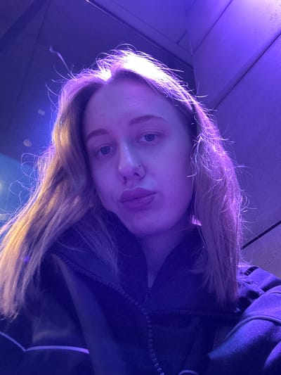 A close-up, portrait-style photograph captures a young woman with fair skin and light brown or blonde hair, illuminated by purple and blue lighting. Her face is slightly tilted, with her eyes open and directed towards the camera. She appears to be wearing a dark jacket. The background is blurred, showing a ceiling with paneling. No specific details indicating the location of Ol'ginskaya, Russia are visible. The image focuses on the woman's face and immediate surroundings.