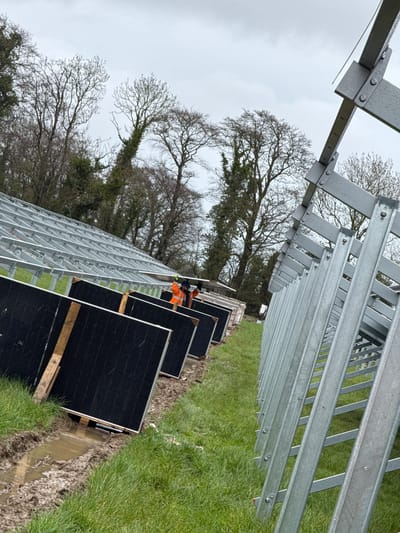 The image captures an outdoor construction scene, identified as a solar farm installation at Baldurgan Hill, Ireland, under an overcast sky. Rows of metallic, galvanized steel support structures for solar panels extend into the background, appearing damp. In the middle ground, two individuals are visible; one worker, wearing a high-visibility orange jacket and a hard hat, is actively handling or positioning a large, black rectangular solar panel. Several other identical black solar panels are propped on wooden pallets or supports on the ground nearby. The terrain features green grass interspersed with significant patches of wet, dark brown mud, including standing puddles. A dense line of bare deciduous trees, some heavily draped in ivy, forms the distant backdrop.