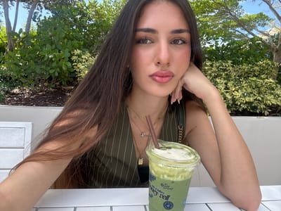 In Hallandale Beach, Florida, USA, a young woman with long, dark brown hair is seated outdoors at a white table, captured in a medium shot. She is leaning her head on her left hand, with her arm resting on the table. The woman is wearing a dark green pinstripe vest with a low-cut neckline, adorned with several necklaces, including a pendant with a cross. A clear plastic cup filled with a green beverage, likely a matcha latte, with a brown straw, sits on the table to her right. Behind her, lush green foliage and trees are visible under a clear blue sky, suggesting a bright, sunny day. The background also shows hints of outdoor seating and a textured white wall.