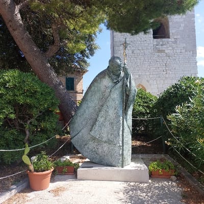 A weathered bronze statue of Pope John Paul II, depicted in robes and holding a crucifix-topped staff, stands centrally on a rectangular, light-colored stone plinth. This monument is located within the landscaped grounds adjacent to the Cattedrale di San Ciriaco in Ancona, Italy.

The statue exhibits a green patina indicative of its material and age. To the left, a large tree with a thick, textured trunk and dense green foliage dominates the upper-left quadrant, partially overhanging the statue. Green bushes flank both sides of the monument. A terracotta pot containing a prickly pear cactus and other green plants is positioned on the ground to the left of the statue, while a smaller terracotta planter with green foliage rests to the right. A low, light-colored rope barrier, supported by dark posts, defines the perimeter of the planted area around the statue.

In the background to the right, a large, light-colored stone building, characteristic of church architecture, features visible masonry and an arched opening. To the left, behind the prominent tree, a section of another building with light-colored walls and a dark green-shuttered window is partially visible. The ground beneath the statue and in the immediate foreground appears to be a light-colored, possibly gravel or compacted earth pathway, with shadows cast by overhead foliage. The scene is illuminated by direct sunlight under a clear blue sky. No individuals are present in the frame.