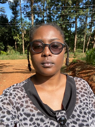 A brown-skinned woman in a spotted blouse and black-rimmed sunglasses poses in a natural setting. Her hair is styled in small braids. Tall trees line the background under a bright blue sky. The ground is a reddish-brown dirt or clay. The woman's expression is serene. The light is bright and casts distinct shadows. The blouse features a dark fabric accent with a large button detail.