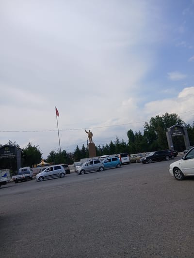 In Bazar-Korgon, Kyrgyzstan, a street scene unfolds under a partly cloudy sky. A statue of a man stands prominently on a pedestal in the middle ground, his arm raised. A red flag flies from a tall pole to the left of the statue. Flanking the statue area are two large archways with inscriptions, the one on the left partially obscured by trees. A road curves through the foreground, populated by several parked and moving vehicles, including a white sedan in the far right, a silver minivan, a black sedan, a blue compact car, and several trucks and other cars. Trees line the background, and a small carousel is visible behind the vehicles. The ground is paved with asphalt.