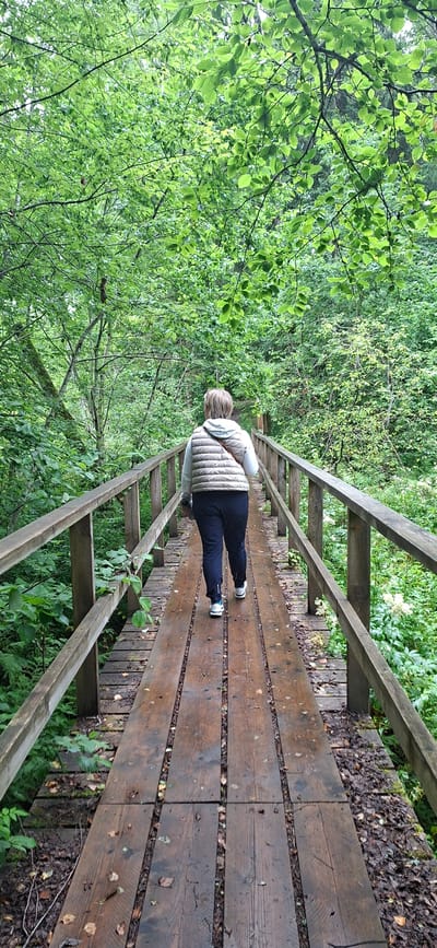 A woman walks away from the camera on a wooden bridge through a forest. The bridge is made of weathered wooden planks with wooden railings on either side. The woman is wearing a light-colored puffer vest over a white shirt and dark pants, and white sneakers. She has short blonde hair and a brown purse slung across her shoulder. The forest is lush and green with dense foliage and trees on both sides of the bridge, suggesting a natural or park setting. The bridge appears to be in the vicinity of Saula, Estonia, based on the context provided.
