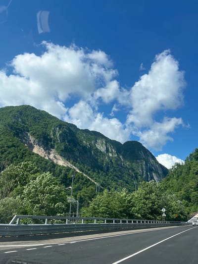 The image shows a landscape view from a road, likely a highway, in Monastyr, Russia. A mountain range dominates the background, with lush green trees covering most of its slopes. A significant scar of lighter-colored earth, indicative of a landslide or erosion, is visible on the side of one of the mountains. The sky is a vibrant blue, dotted with fluffy white clouds. In the foreground, a paved road stretches into the distance, with a metal guardrail separating it from the surrounding vegetation. There are no people clearly visible in the image. The environment suggests a natural, mountainous region.