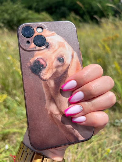 A close-up shot shows a person's right hand holding a smartphone with a case featuring a printed image of a golden retriever's face. The phone is positioned in front of a blurry background of green grass and foliage, suggesting an outdoor setting. The person's nails are manicured with a French tip design, featuring pink and white polish with a subtle glitter accent. A gold-colored wristwatch is visible on the person's wrist. The image appears to have been taken in Karniowice, Poland, based on contextual information.