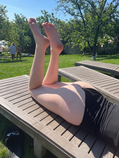 The image shows a person lying on a wooden lounge chair outdoors on a sunny day. The subject is wearing black shorts and a black top. Their legs and feet are visible, with the feet in the foreground. The scene is set in a grassy area with trees and a stone wall in the background. The sun is shining brightly, casting shadows on the lounge chair and the subject's legs. In the background, other people can be seen walking on the grass. The overall impression is one of a relaxed, summery atmosphere. The time of day appears to be afternoon, given the angle of the shadows and the brightness of the sunlight. There are no visible emotions expressed as the focus is on the legs and feet. There is visible text on a black sandal in the lower right corner of the image which reads "FILA SINCE 1911".