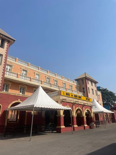 A tall, multi-story building is depicted with a combination of red brick and peach-colored textured facade. A large yellow sign vertically displays the text "I WR H RAI WA ATIO." The building features arched openings and decorative railings on some floors. White tents are positioned on the right side of the image, adjacent to the building. The ground is paved, and the sky is clear blue. Based on the signage, the location is potentially in Anajpur, India.