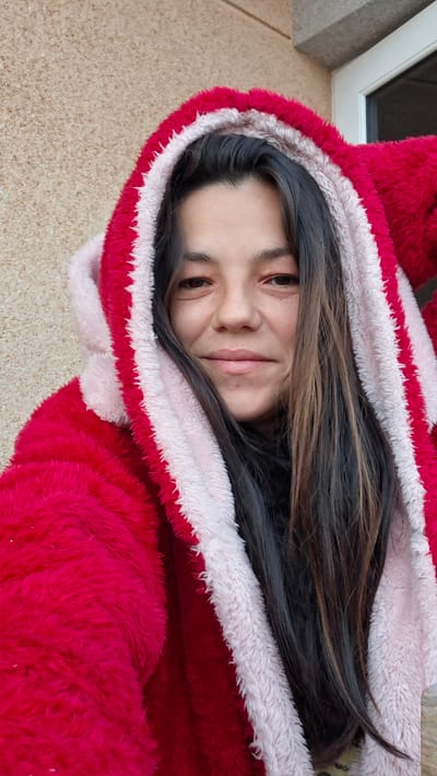 A woman is depicted in a medium close-up, facing forward with a slight smile and direct gaze towards the viewer. She has long, dark brown hair, which falls over her shoulders and partially within the hood of her garment. She is wearing a fluffy, hooded garment that is predominantly bright red, with the hood's interior lining and trim appearing in a light pink or white color. The garment's texture is soft and plush. The background consists of a light brown, coarse-textured wall on the left side, with a segment of a white-framed window or door visible in the upper right quadrant. The scene is located in Granadilla de Abona, Spain.