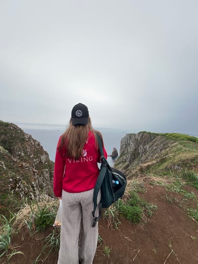 The image depicts a person standing on a cliff overlooking the sea in Russkiy Island, Russia. The person wears a black cap, a red sweater with the word "VIKING" on the back, and plaid pants. A backpack rests on their back. The landscape features rocky cliffs, grassy terrain, and a foggy or overcast sky. The sea is visible below, with some rocky formations emerging from the water.