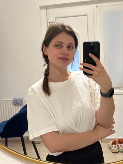 A young woman takes a selfie in a bathroom mirror. She's wearing a white t-shirt, a dark scrunchie in her hair, and a green smartwatch. She's holding a black smartphone. The bathroom is white, with a window and radiator visible in the reflection. There is no indication of Kőröshegy, Hungary in the provided image.