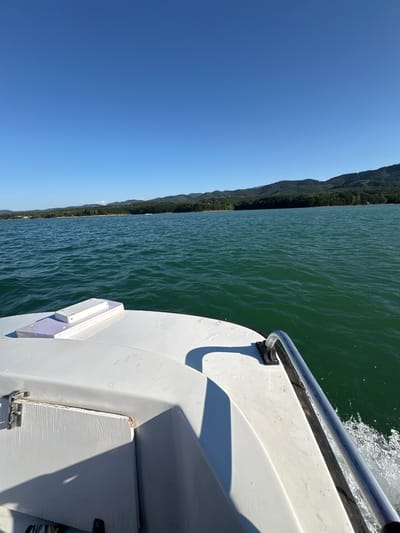The image shows a view from the bow of a white boat moving through a body of water. The water is a deep green color with ripples on the surface. In the distance, there is a shoreline covered in dense green trees, followed by rolling hills under a clear blue sky. The foreground of the image focuses on the boat's deck, showing white fiberglass surfaces, a metal railing, and some white equipment mounted on the deck. There are no people visible in the image. The scene appears to be a serene day on the water, likely in the vicinity of Golyama Zhelyazna, Bulgaria, a location known for its natural beauty and reservoirs.
