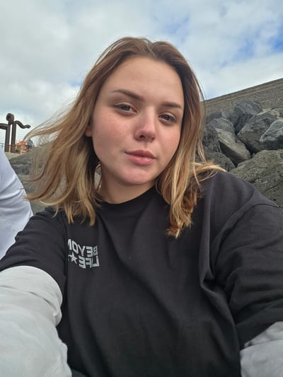 This is a selfie of a young woman with blonde hair, taken outdoors. She is wearing a black long-sleeved shirt over a white long-sleeved shirt. On her black shirt, there is a silver graphic that appears to be text and a star. The woman is looking towards the camera, with a neutral expression.

Behind her, to her left, is a rocky cliff face and a cloudy sky. To her right and lower in the frame, part of what appears to be a metal structure with ornate scrollwork is visible, along with a sliver of blue sky peeking through the clouds. The overall lighting suggests it is daytime, possibly overcast. The rocks and the ocean in the background (implied by the Puerto De La Cruz location context) suggest a coastal setting. The scene appears to be relaxed and casual, possibly a tourist or recreational outing.