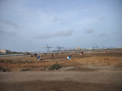 The image shows a group of people playing a game of soccer on a sandy beach in Port Sudan, Sudan. The beach is sparsely vegetated and is located near the waterfront.  In the background, the Port Sudan harbor is visible, featuring several large cranes and ships. The sky is overcast with a light blue color.  The people playing soccer are all wearing casual clothing. Several people are sitting on the beach, watching the game.