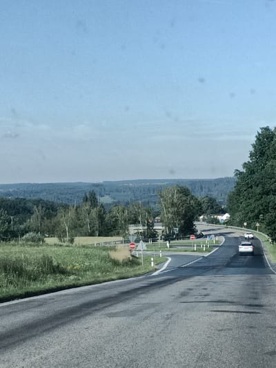 The image captures a scenic view of a road winding through a rural landscape in Šternov, Czechia. The scene unfolds on a clear, bright day, suggesting it's likely daytime with good weather conditions. The road itself, paved with asphalt, dominates the foreground and curves into the distance, leading towards a gentle hill.
Along the sides of the road, lush green grass and vegetation are abundant, with some patches of dry, taller grass adding texture. In the mid-ground, several cars are visible. Two white cars are driving up the road, one directly behind the other. Further ahead, another white car is also traveling in the same direction. A few red and white traffic signs, common in European countries, are placed along the road's edge, indicating traffic regulations.
The background is a panorama of rolling hills covered in dense, dark green forests. The distant horizon is softened by a hazy blue sky, typical of a clear day. A few sparse clouds are also present in the sky. The overall impression is one of peacefulness and natural beauty, with the road serving as a gentle pathway through the expansive countryside. No people are explicitly visible in the frame, but the presence of cars suggests ongoing travel. The location is characterized by its natural, untamed scenery, hinting at a rural or semi-rural setting.
