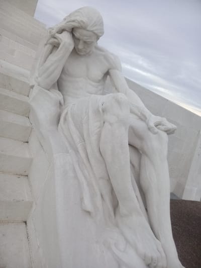 A white marble sculpture of a reclining male figure is depicted. He appears to be in a state of sorrow or despair, with his head resting on his hand. His body is draped in fabric. The sculpture rests on a stone base with visible seams. The background consists of cloudy sky and architectural elements, suggesting the sculpture is part of a larger monument.