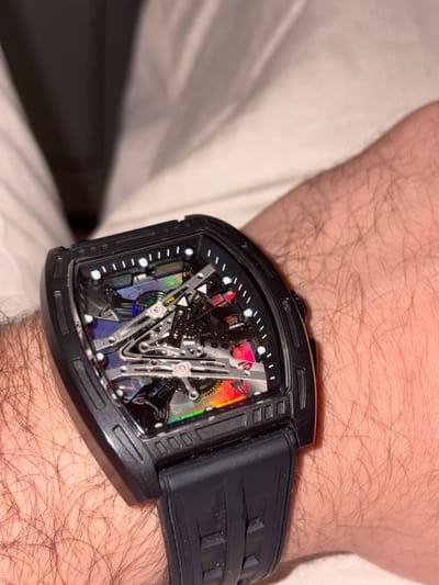 A close-up shot reveals a hairy arm adorned with a black, angular watch featuring a visible mechanical movement. The watch's intricate gears and components are on display beneath the crystal. The watch band is textured and also black. The arm rests against a light-colored, possibly white, fabric. The location is not discernible from the image.