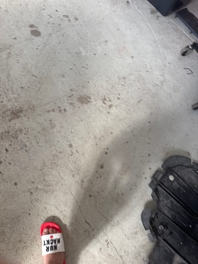 In Schkopau, Germany, a person is wearing a white slide with red trim on their left foot. The slide has black lettering that reads "NUR NACKT" upside down. The person is standing on a concrete floor that has several dark stains and a visible crack. To the right of the foot, a black object with metal components is present on the floor, casting a shadow. The environment appears to be indoors, possibly a garage or workshop, given the concrete floor and the presence of mechanical-looking parts.