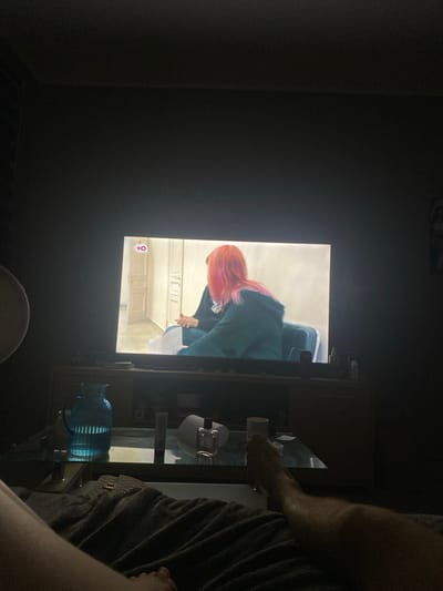The image shows a dimly lit living room with a television displaying a program featuring two individuals. One individual on the television has bright pink hair and is wearing a dark green hooded sweatshirt. The television is situated on a wooden entertainment stand. On a glass coffee table in front of the stand, there is a blue glass pitcher, a roll of paper, a bottle of lotion, and a small glass bottle. The lower portion of the image shows a person's legs covered by a blanket. The overall setting suggests a relaxed evening at home. There is no information in the image to suggest a specific location in Tartu, Estonia.