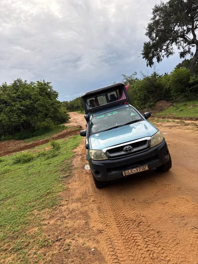 A light blue Toyota pickup truck parks on a dirt road in a rural setting. One door is open, revealing a glimpse of the vehicle's interior and a person inside. Lush green vegetation borders the road on one side, while the other is mostly dirt and sand with visible tire tracks. Trees and a cloudy sky fill the background. The license plate on the front of the truck is partially visible, displaying "LE=9707."