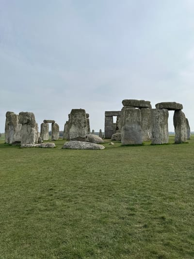 The image displays Stonehenge, a prehistoric monument located near Amesbury, Wiltshire, United Kingdom. The scene features a series of massive grey sarsen stones and smaller bluestones arranged in an elliptical or circular configuration. Many stones stand upright, some forming recognizable trilithons with horizontal lintel stones atop two vertical uprights, while others are fallen or partially buried. The stones exhibit natural weathering and textural variations across their surfaces. The ground in the foreground and surrounding the monument is covered by short, vibrant green grass, indicative of a field or plain. The sky above is overcast with a uniform light grey cloud cover. No individuals, animals, or specific actions are visible within the frame. The horizon in the distance shows a subtle line of trees under the grey sky.