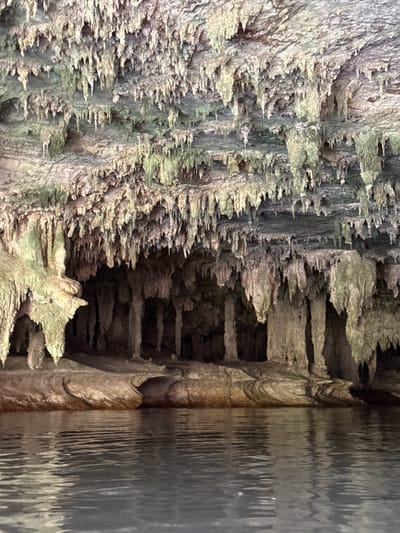 This media file depicts an underground cave formation, likely a limestone cave, characterized by numerous stalactites and stalagmites. The cave walls are rough and textured, with formations hanging from the ceiling and rising from the floor. Some of these formations are covered in a light green mineral deposit, possibly algae or moss, suggesting a damp environment. In the right portion of the image, a body of water is visible, reflecting the cave formations and creating rippling patterns on its surface. The lighting is dim, typical of an indoor cave environment, with some areas illuminated by an unseen light source. The overall scene evokes a sense of natural wonder and geological time. There are no people or text visible in the image. The location is given as Ao Luek, Thailand, which is known for its limestone karst landscapes and caves.