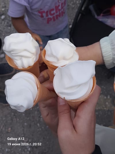 Four ice cream cones, topped with soft-serve ice cream, are held by multiple hands. In the background, a child wearing a purple t-shirt with the words "TICKLE PINK" is visible. The scene is outdoors, with a grey, textured surface that appears to be pavement. The image contains a timestamp "Galaxy A12 19 июня 2026 г. 20:52". The location is identified as Kara-Köl City, Kyrgyzstan.