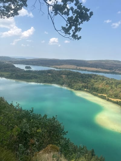 The image is a landscape view from a high vantage point overlooking multiple lakes and surrounding land. In the foreground, lush green foliage and dry grasses are visible on a slope. The main subject is the series of lakes, characterized by their striking turquoise and lighter, sandy-colored waters. The lakes are bordered by dense green forests and rolling hills in the background, with some fields and a few scattered buildings. The sky is a bright blue with scattered white clouds. The perspective suggests the photograph was taken on a clear, sunny day from a clifftop or elevated viewpoint in the vicinity of La Chaux-du-Dombief, France, known for its natural beauty and lakes. There are no people or animals visible in the frame.