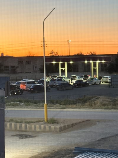 At sunset, a gas station in Zhanaozen, Kazakhstan, is busy with multiple cars queuing at the pumps. The sky is a vibrant gradient of orange and pale blue. The gas station canopy is illuminated by bright lights, casting a glow on the vehicles below. A tall streetlight stands prominently in the foreground, its pole partially obscured by a yellow segment. The scene depicts a common urban activity, with the natural beauty of the sunset providing a striking backdrop. The area appears to be a typical roadside development, with concrete barriers and low vegetation surrounding the gas station.