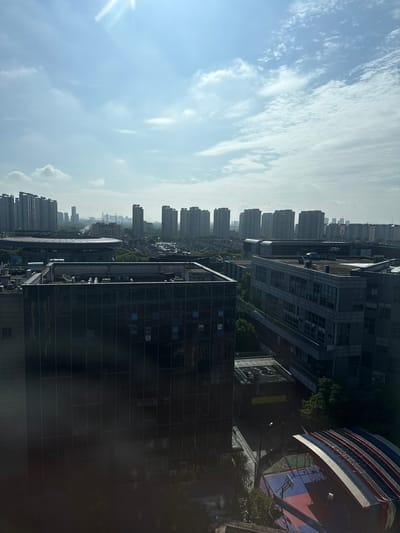 The image captures an urban cityscape under a bright, partially cloudy sky in Minhang District, China. In the foreground, the dark, reflective facade of a modern office building dominates the lower left portion of the frame. The building features numerous windows that reflect the sky and surrounding environment. To the right of this building, another multi-story office complex with a grey, metallic exterior and visible window panes is situated. A smaller building with a dark roof and some visible air conditioning units is nestled between these larger structures.

Further into the background, a circular building with a distinct tiered roof structure and a glass enclosure is visible, surrounded by lush green trees. Beyond this, a dense arrangement of residential high-rise buildings stretches across the horizon, interspersed with lower-rise structures and more greenery. The sky is a pale blue, with streaks of white clouds and a bright sunbeam emanating from the upper left corner. In the lower right of the image, a section of an outdoor recreational area is visible, featuring a red and blue court marked for basketball and a covered seating area with red, white, and blue striped awnings. No people are clearly visible in the image. The scene depicts a typical day in a developed urban area.