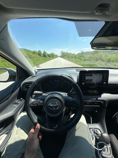 The image shows a view from inside a car driving on a road near Dolna Beshovitsa, Bulgaria. The dashboard and steering wheel are prominent. A navigation system displays a route. The driver is holding the steering wheel with their right hand. The exterior view shows a road and a landscape with trees and hills under a clear sky.