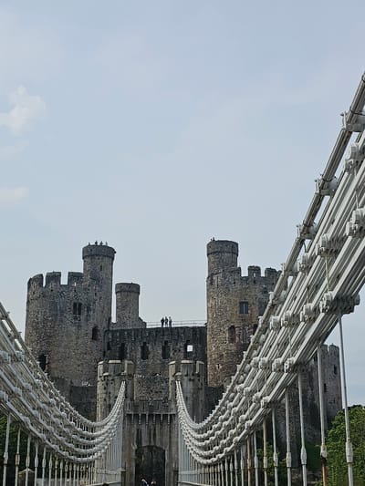 The image shows a view of Conwy Castle, a medieval fortification in Conwy, Wales, United Kingdom. The castle is constructed of gray stone and features multiple towers and battlements. It appears to be viewed through the support structure of a nearby bridge. The sky is visible in the background, mostly clear with light clouds. The image is taken from an angle, likely from below the bridge structure, providing a unique perspective of the castle.