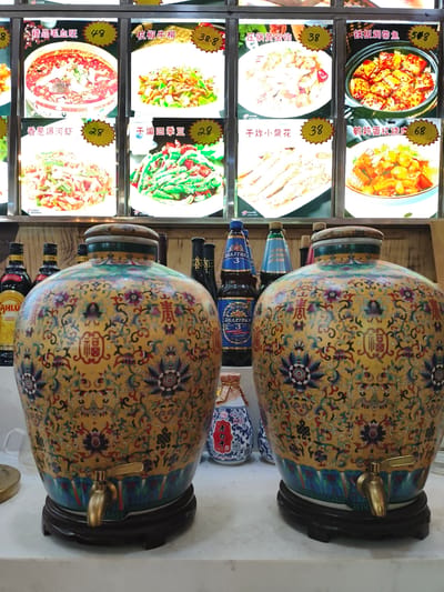 The image captures a close-up view of two large, ornate ceramic dispensers, likely for beverages, situated on a counter in what appears to be a restaurant or food establishment. These dispensers are decorated with intricate patterns featuring floral motifs and Chinese characters, primarily in yellow, blue, red, and green. Each dispenser has a golden spigot at its base and rests on a dark wooden stand.

In the background, a menu board displays images of various Chinese dishes with their prices. To the left and right of the dispensers, several bottles are visible, including some with labels that suggest alcoholic beverages like coffee liqueur and beer. The lighting in the background is bright, illuminating the menu items.

The scene suggests a setting where customers can serve themselves drinks from the dispensers while perusing the food options. The overall impression is one of a functional yet decorative establishment, possibly a casual eatery in Jixi, China, given the presence of Chinese script on the menu and the style of the food displayed. There are no people visible in the image.