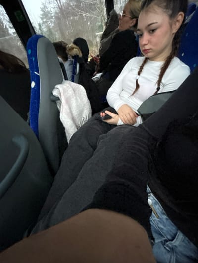 A teenage girl with braided hair sits on a bus. She wears a white long-sleeved shirt and a grey seatbelt. Next to her, someone wears black leggings. A white towel sits atop a blue bus seat. In the background, a woman with blonde hair and glasses sits near a window. Outside, snow-covered trees are visible. The bus interior appears dimly lit, adding a slightly gloomy atmosphere.