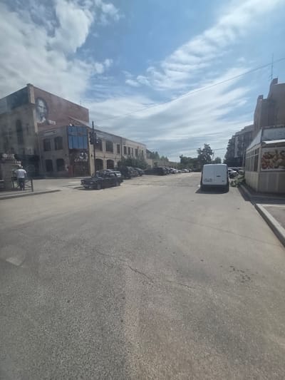 The image shows a street scene in the city of Gyumri, Armenia, near the Border Gyumri-Akhuryan. A black car is parked on the right side of the road. In the background, a white van is parked, and several other vehicles are visible. Buildings line both sides of the street. One building on the left has a large mural of a person's face painted on its side. The sky is cloudy, with patches of blue visible. The overall impression is of a somewhat quiet urban environment on a partly cloudy day.