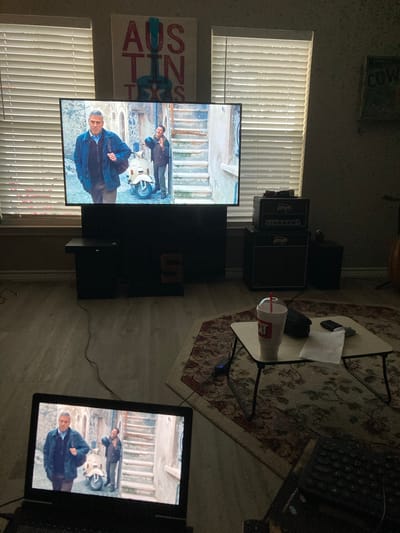 The image depicts an indoor living room scene, likely in Round Rock, Texas, as indicated by a piece of wall art with "AUSTIN TEXAS" visible. Two screens are showing a movie with actors George Clooney and another individual, possibly from the film "The Tourist". The main television is a large flatscreen displaying the movie, positioned on a dark entertainment stand. To the right of the television are two stacked guitar amplifiers and a smaller black speaker. In the foreground, a laptop computer is open, also displaying the same movie scene. A small foldable table sits on an oriental rug, holding a white cup with a red straw, a black object, and a crumpled white napkin. In the background, horizontal blinds cover a window, and another piece of wall art with the word "COW" is visible. The room is dimly lit, suggesting it is evening or the blinds are closed. The flooring appears to be light-colored laminate or wood.