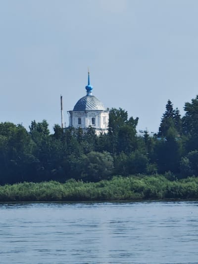 A white church with a blue dome and a cross is visible across a wide river. The church has a classical architectural style with arched windows. It is surrounded by lush green trees, and a tall, slender structure, possibly a chimney or antenna, is situated to its left. The sky is a clear, pale blue. The foreground features the tranquil surface of the river, with gentle ripples reflecting the light. The scene suggests a peaceful, possibly historic, location. The church is likely the Church of St. Nicholas the Wonderworker, located in Yeniseysk, Russia.
