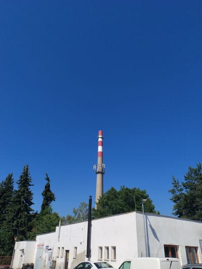 This is a photograph of a power plant chimney. The chimney is tall and cylindrical, with red and white stripes. It is set against a clear blue sky. To the right of the chimney is a white building with several windows. There are trees in the foreground and background. The photograph appears to have been taken during the day, as the sun is shining. The scene is likely a power plant in Písek, Czechia. There is text visible on the building which reads "TOWCE TURES".