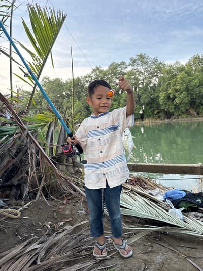 A young boy stands outdoors, holding a fishing rod and a small fish he has caught. He is wearing a striped shirt and jeans, and is barefoot in flip-flops. The boy is standing on a dirt path, with palm leaves scattered around him. In the background, there is a body of water, likely a river or lake, and lush green trees. The sky is cloudy, suggesting it may be late afternoon or early morning. The overall scene depicts a peaceful, rural setting. The location is identified as Grong Grong Capa, Indonesia, a place known for its natural beauty and opportunities for outdoor recreation.