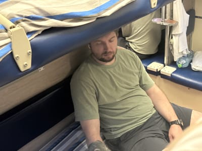 A man with short brown hair and a beard is sleeping with his eyes closed. He is wearing a green t-shirt and dark shorts, and has a black watch on his left wrist. He is lying on a bunk bed with a blue mattress and a striped blanket. There is another bunk bed above him, and a water bottle and some white fabric are visible on a shelf. The scene appears to be inside a train compartment, possibly on the Trans-Siberian Railway, given the location of Байлянгар, Russia.