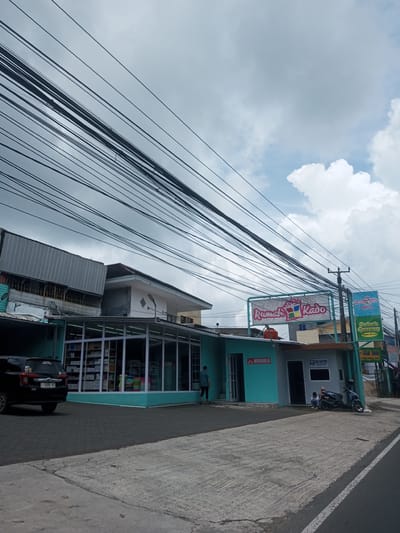 The image shows the exterior of a teal-colored store called "Rumah Kado", with a sign of the same name above the entrance, located on a street. A black SUV is parked in front of the store. A person is walking towards the entrance of the building, and a motorcycle is parked next to a guard house. A sign for "Bebek Goreng Mang Aceng" can be seen. A dense network of overhead power lines dominates the sky. The location is in a city environment. The text on a license plate reads "F 1590 OT". The image does not give any indication of location in Laos.
