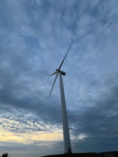 A low-angle shot captures a towering white wind turbine against a dramatic, cloudy sky. The turbine's long, slender blades are angled upwards and to the left, with the rotor hub visible at their intersection. The sky is a mosaic of dark, brooding clouds interspersed with patches of lighter, illuminated clouds, suggesting either dawn or dusk. A strip of lighter sky with yellow and orange hues is visible near the horizon line. The base of the turbine is grounded on a slightly elevated, dark terrain, with a hint of vegetation or low bushes. A small structure, possibly a maintenance shed or entrance, is visible at the base of the turbine tower. In the distance, a faint horizontal line suggests the presence of land or structures, potentially a coastline or the edge of the landscape in Nasva alevik, Estonia. The overall atmosphere is one of natural power and a sense of vastness.