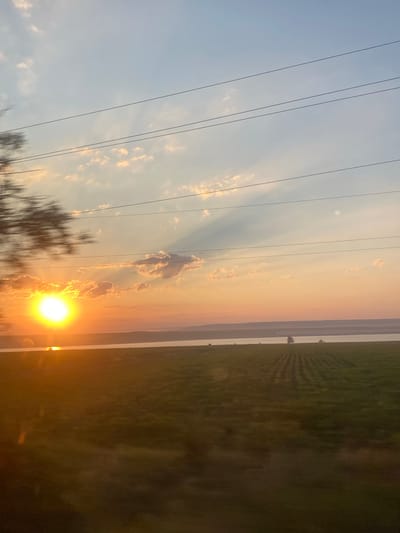 The image captures a sunset scene in Balabanu, Moldova. The sun, a bright orb of light, is low on the horizon, casting a warm, golden glow across the sky and reflecting on the surface of a body of water. Wispy clouds are scattered across the sky, illuminated by the setting sun. In the foreground, a field of green crops is visible, with rows clearly defined by shadows. A solitary tractor is positioned in the field, near the edge of the water. In the distance, a hazy landscape of rolling hills can be seen. Several power lines are strung across the sky, partially obscuring the view. The overall impression is one of a serene, rural landscape at dusk.