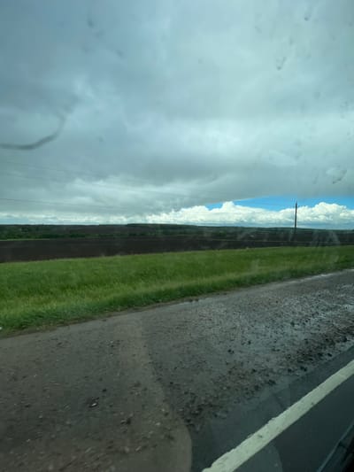 The image shows a view from a vehicle driving on a road in what appears to be a rural area. To the left is a view of a cloudy sky with some clear, blue sky visible in the distance. Along the horizon is a line of cumulus clouds. There is a utility pole in the middle distance, and power lines run across the scene. Adjacent to the road is a grassy area, and further beyond that is a strip of dark soil. The road surface is wet and asphalt, with a white lane marking visible on the right edge. The image is tilted, and the view appears to be from inside the vehicle, as evidenced by the watermarks on the glass. There is no indication this picture was taken in Кокино, Russia.
