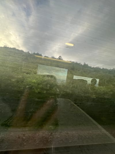The image shows a reflection on a surface, possibly a window, of a landscape and two rectangular objects, one on the left and one on the right, reflecting portions of the landscape and a sky with clouds. The landscape appears to be a green, hilly area with trees and foliage. The sky is overcast with some brighter areas. There are also reflections of what appear to be two human-like figures in the foreground, with the reflection of a face visible in the rectangular object on the right. The overall impression is of a distorted or layered reality, with the viewer seeing both the external environment and reflections of what is inside or on the surface. The location is identified as being in the city of Dover, United Kingdom, though no specific landmarks or features within Dover are identifiable from the image.