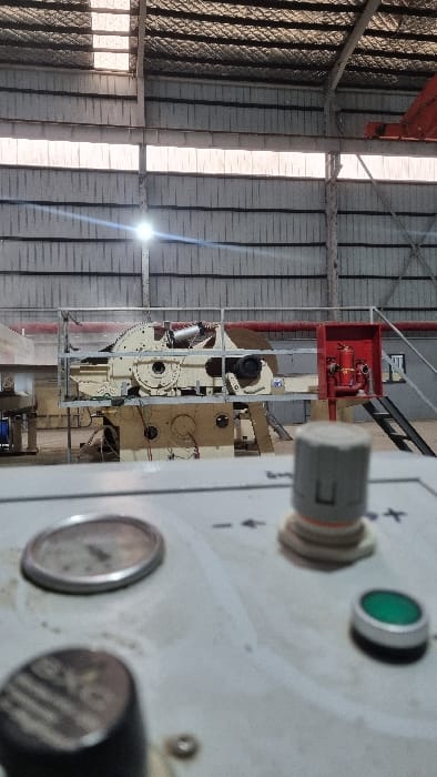 The image presents an interior view of an industrial facility, likely a factory or processing plant, situated in Chapyk, Uzbekistan. The foreground is occupied by a blurred, light-colored control panel featuring a grey rotary knob marked with a vertical line and a horizontal base, a circular metallic gauge, and a green push-button. Textual markers including '-', '+', and 'x' are visible near the knob.
In the midground, a large, cream-colored industrial machine is prominently displayed, resting on an elevated platform. This machinery incorporates exposed mechanical components such as gears, shafts, and a prominent disc-shaped element on its right side. Electrical wiring and hoses are connected to various parts of the machine. To the immediate right, a red fire suppression system, including pipes, valves, and a hose reel, is mounted within a red cabinet on a grey wall. A dark metal staircase descends behind this fire station.
The background reveals a corrugated metal ceiling and walls, characteristic of a warehouse or factory structure. Structural beams are visible overhead. Natural light enters through high windows or skylights, creating a bright glare in the upper-left portion of the image. The floor appears to be concrete. No personnel are present in the scene.