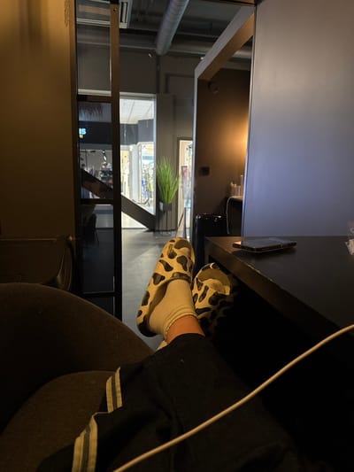 This image captures a person's lower body at rest within a modern commercial interior, specifically located within or adjacent to Ålesund Storsenter in Ålesund, Norway.

The foreground displays the lower legs and feet of an individual, seated in a dark upholstered chair, with their right foot elevated. The person is wearing dark pants with a white stripe along the outer seam, white socks, and black-and-white cow-print patterned slide sandals. A dark rectangular table is positioned in front of the individual, supporting a dark-colored smartphone resting face-down. A white cable is visible, extending from the bottom right of the frame, crossing the person's leg towards the table.

The background reveals a contemporary indoor environment with dark walls and exposed silver-colored industrial-style ductwork on the ceiling. To the right of the table, a dark waste bin is partially visible. Further back and to the right, a warm, ambient light source illuminates a section of a dark wall. In the center and left background, a brightly lit shopping corridor is visible through a dark-framed partition or doorway, with reflections from storefronts. Text, including "CUBES," is discernible in reflections or through glass, indicating a retail space consistent with Ålesund Storsenter. A tall, slender artificial plant with green foliage in a dark planter stands in the central background. The scene suggests a lounge or waiting area within a shopping complex.